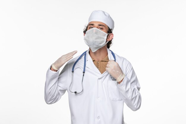 Front view male doctor in medical suit wearing sterile mask as a protection from covid on light-white desk virus coronavirus pandemic illness health