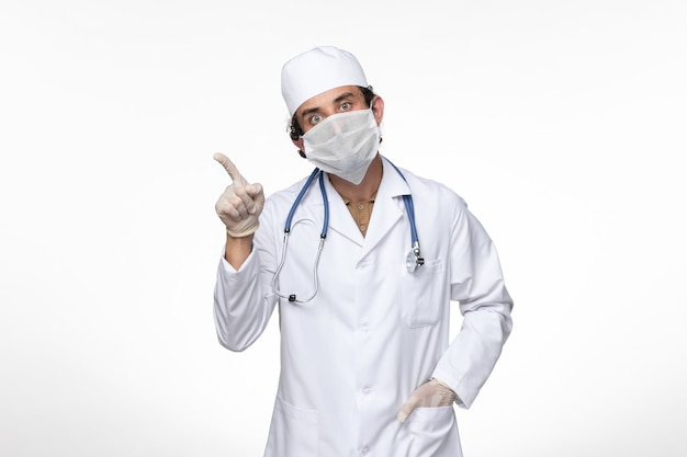 Front view male doctor in medical suit wearing sterile mask as a protection from covid- on light-white wall virus illness covid pandemic disease