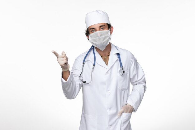 Front view male doctor in medical suit wearing sterile mask as a protection from covid- posing on white wall virus illness covid pandemic disease