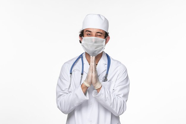 Front view male doctor in medical suit wearing sterile mask as a protection from covid praying on white wall virus coronavirus pandemic illness disease