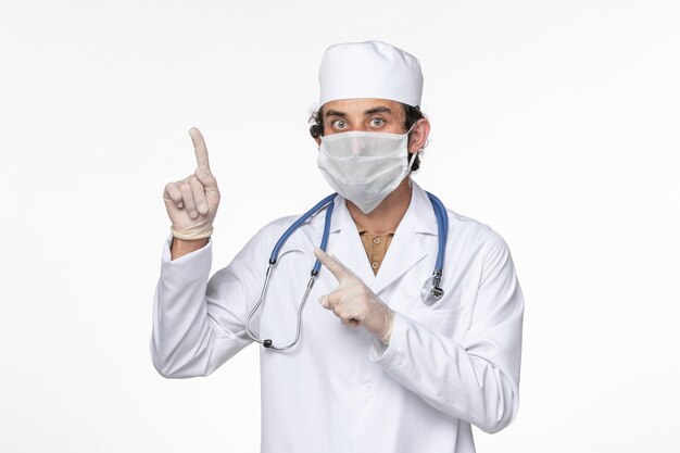 Front view male doctor in medical suit wearing sterile mask as a protection from covid- raising his finger on white wall virus coronavirus pandemic disease