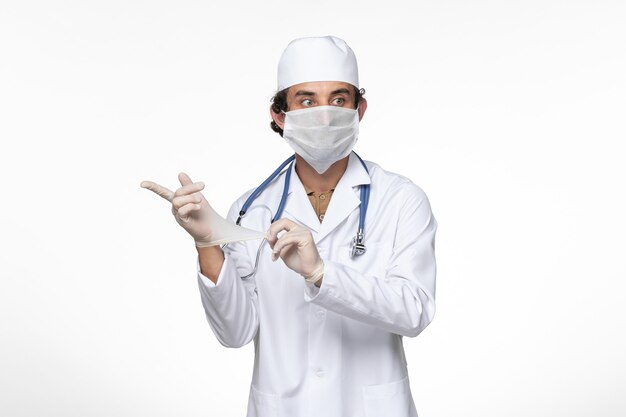 Front view male doctor in medical suit wearing sterile mask as a protection from covid- wearing gloves on white wall virus illness covid pandemic disease