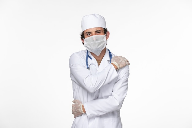 Medico maschio di vista frontale in vestito medico che indossa maschera sterile come protezione da covid sulla salute pandemica del coronavirus del virus della spruzzata della parete bianca