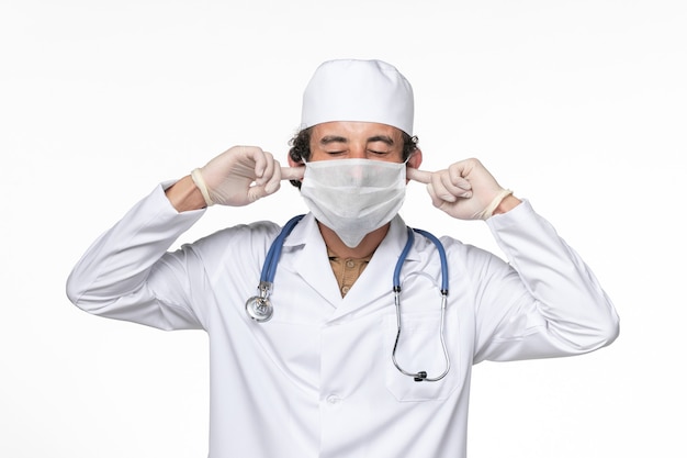 Front view male doctor in medical suit with mask as a protection from covid- shutting his ears on white wall coronavirus virus pandemic medicine