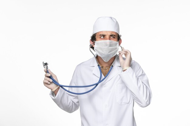 Front view male doctor in medical suit with mask as a protection from covid- using a stethoscope on the white desk splash coronavirus virus pandemic