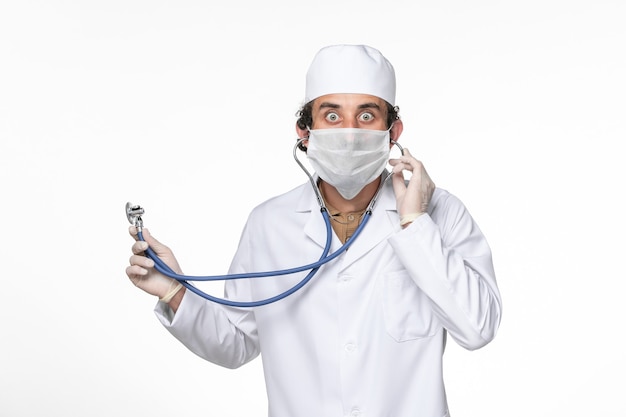 Front view male doctor in medical suit with mask as a protection from covid- using a stethoscope on white desk splash coronavirus virus pandemic