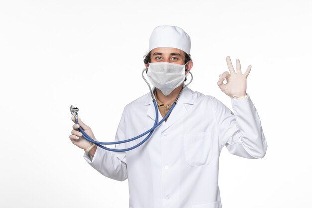 Front view male doctor in medical suit with mask as a protection from covid- using stethoscope on the white wall splash coronavirus virus pandemic