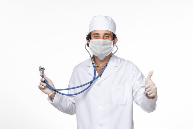 Front view male doctor in medical suit with mask as a protection from covid- using stethoscope on white wall splash coronavirus virus pandemic