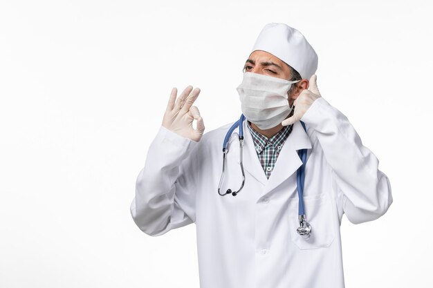 Front view male doctor in medical suit with mask due to covid- on the white desk