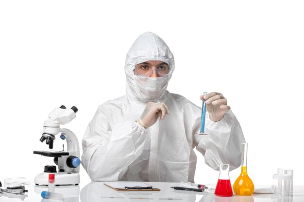 白い机の上に青い溶液と一緒にフラスコを保持しているため、マスク付きの防護服を着た男性医師の正面図