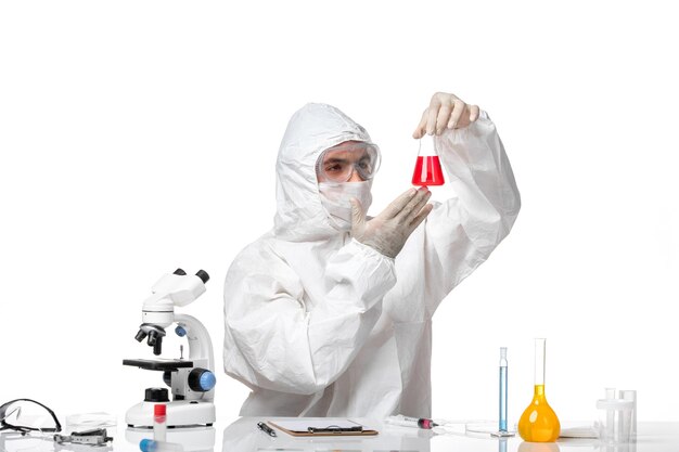 Front view male doctor in protective suit with mask due to covid holding flask with red solution on white desk