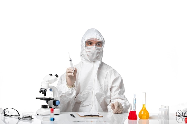 Вид спереди мужчина-врач в защитном костюме с маской из-за covid, держащего инъекцию на белом столе