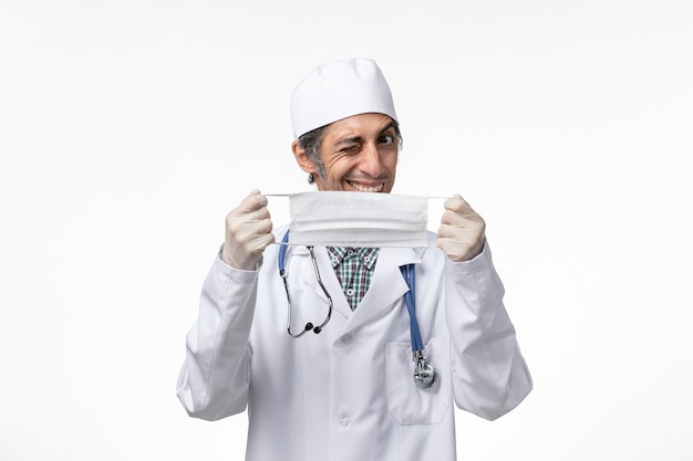 Front view male doctor in white medical suit due to coronavirus holding mask on white surface