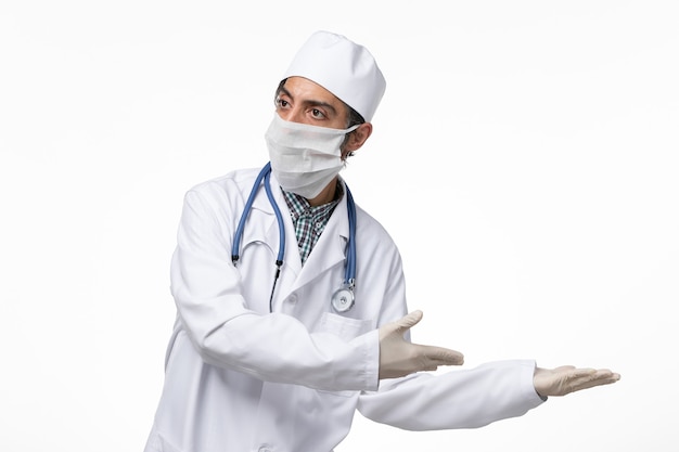 Front view male doctor in white medical suit and mask due to coronavirus on white desk