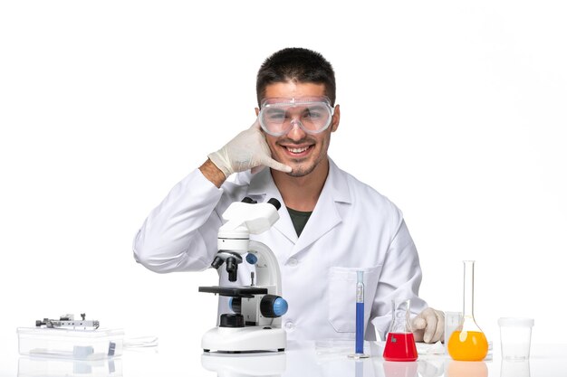 Front view male doctor in white medical suit sitting with solutions and smiling on white space