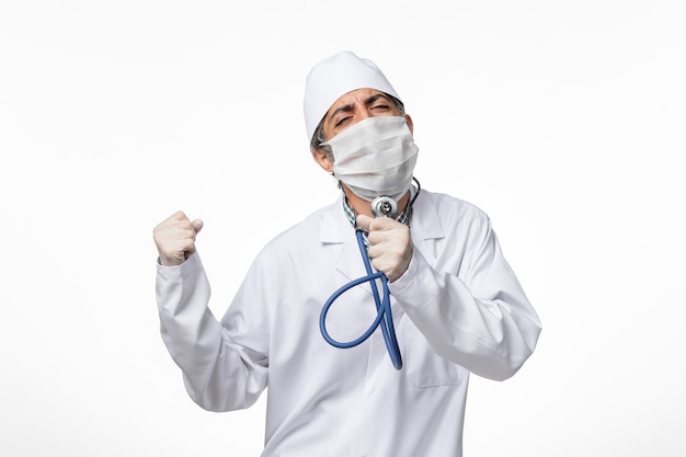 Вид спереди мужчина-врач в белом медицинском костюме в маске из-за коронавируса держит стетоскоп и поет на белом столе