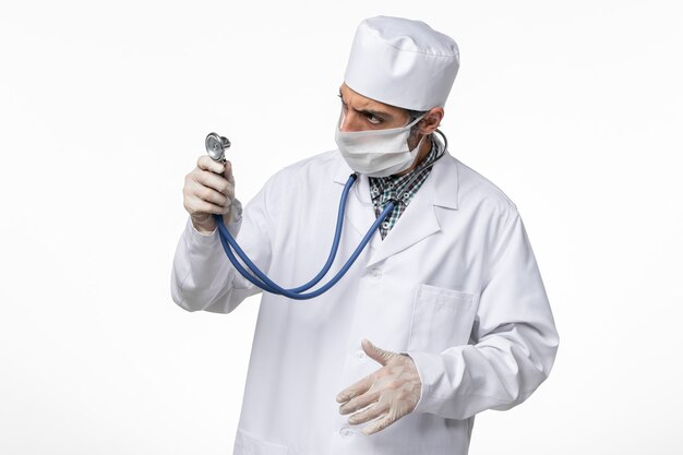 Front view male doctor in white medical suit wearing mask due to coronavirus holding stethoscope on a white desk