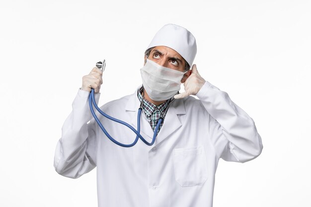 Вид спереди мужчина-врач в белом медицинском костюме в маске из-за коронавируса, держащий стетоскоп на белом столе