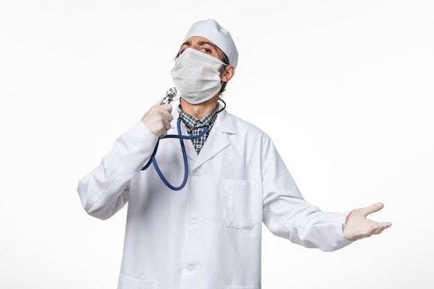 Free photo front view male doctor in white medical suit wearing mask due to coronavirus holding stethoscope on white desk