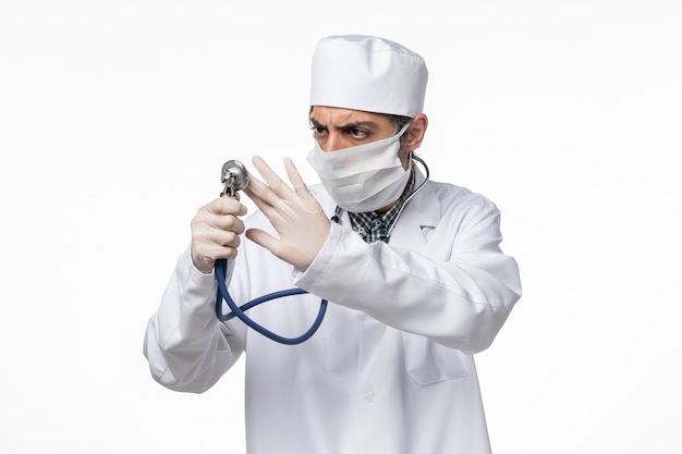 白い表面に聴診器を保持しているコロナウイルスのためにマスクを身に着けている白い医療スーツの正面図男性医師