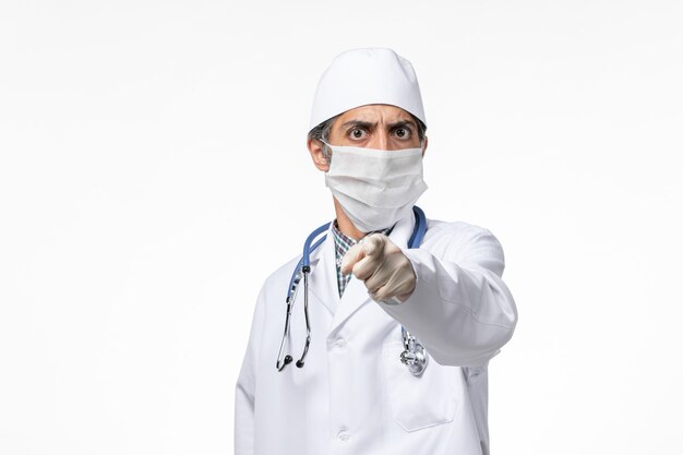 Front view male doctor in white medical suit wearing mask due to covid on a light white desk