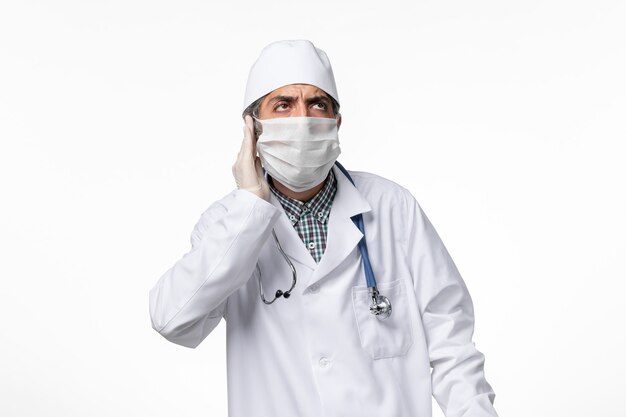 Front view male doctor in white medical suit with mask due to coronavirus on light white surface