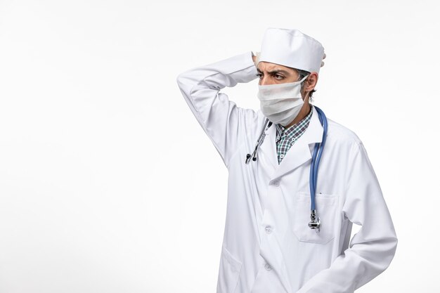 Front view male doctor in white medical suit with mask due to coronavirus thinking on white surface