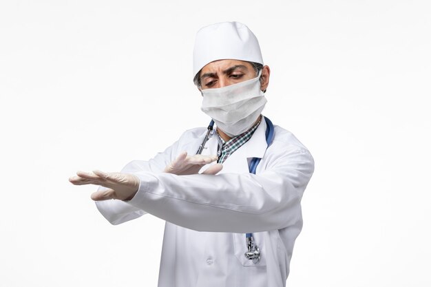 Front view male doctor in white medical suit with mask due to coronavirus on white desk