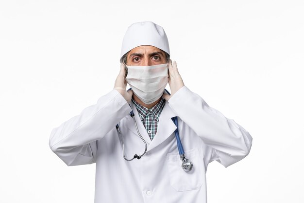 Front view male doctor in white medical suit with mask due to coronavirus on a white surface