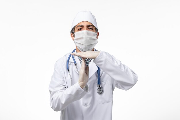 Front view male doctor in white medical suit with mask due to covid on light white surface