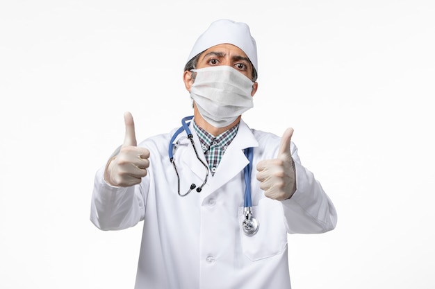 Front view male doctor in white medical suit with mask due to covid showing like signs on white surface