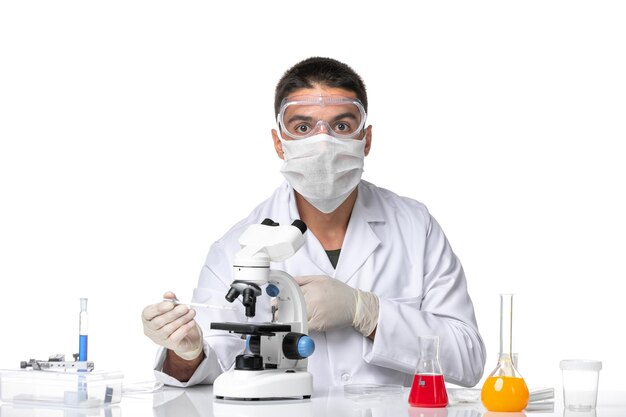 Вид спереди мужчина-врач в белом медицинском костюме с маской из-за covid с помощью микроскопа на светлом белом пространстве