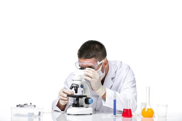 Front view male doctor in white medical suit with mask due to covid using microscope on white desk