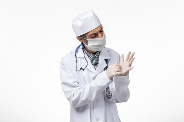 Front view male doctor in white medical suit with mask due to covid wearing gloves on a white surface