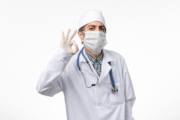 Front view male doctor in white medical suit with mask due to covid on white surface