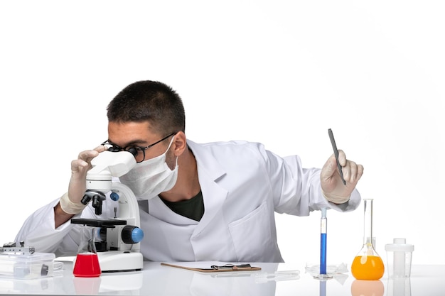 Front view male doctor in white medical suit with mask due to covid working with microscope on white space