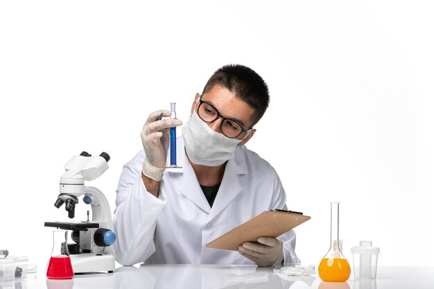 Front view male doctor in white medical suit with mask due to covid writing notes on a white desk