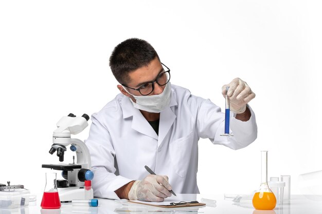 Front view male doctor in white medical suit and with mask working with solution on white desk