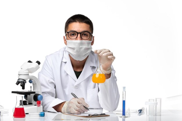 Front view male doctor in white medical suit and with mask working with solutions on white desk