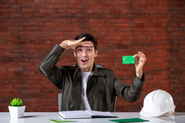 front view male engineer sitting behind his working place and holding credit card builder money plan corporate business property agenda