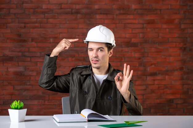 Front view male engineer sitting behind his working place in white helmet document business contractor corporate plan property agenda builder