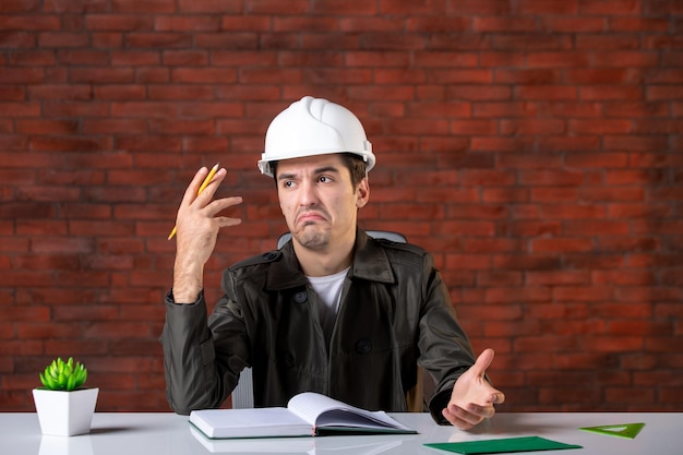 Free photo front view male engineer sitting behind his working place in white helmet document business corporate property job builder contractor plan