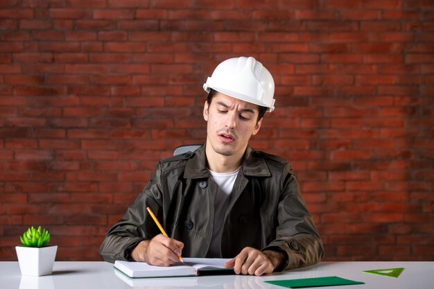 Front view male engineer sitting behind his working place in white helmet document plan business corporate job agenda builder contractor property