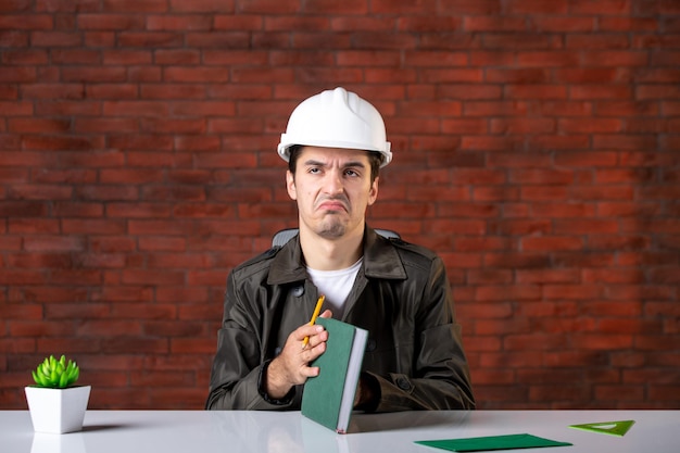 Free photo front view male engineer sitting behind his working place in white helmet document plan business corporate job builder contractor agenda