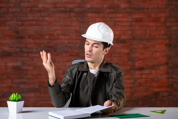 Вид спереди инженер-мужчина, сидящий за своим рабочим местом в белом шлеме, написание заметок, план документа, бизнес-подрядчик, собственность, повестка дня, корпоративный