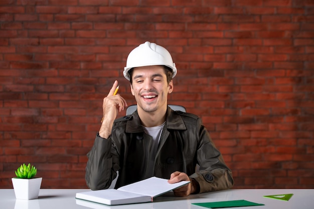 Front view male engineer sitting behind his working place in white helmet