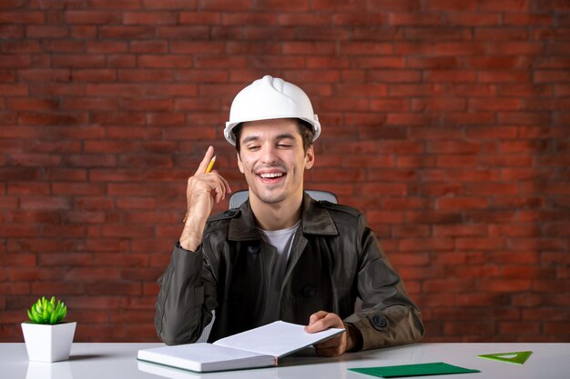 Front view male engineer sitting behind his working place in white helmet