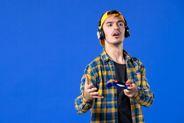파란색 벽에서 게임패드와 헤드폰으로 비디오 게임을 하는 남성 게이머의 전면