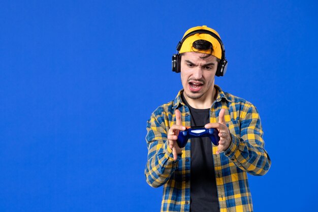 Front view of male gamer with gamepad and headphones playing video game on blue wall