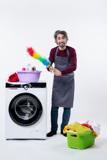 Front view male housekeeper holding feather duster standing near washing machine on white wall